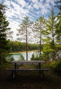 Picnic table in forest with lake and cloudy sky in background.