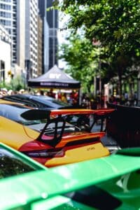 Street scene with colorful GT3 RS car, rear spoiler, tent, tall buildings, and people.