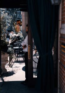 Shaded Doorway Scene with Masked Individual Person in white shirt and mask partially visible in doorway, with outdoor seating and plants.