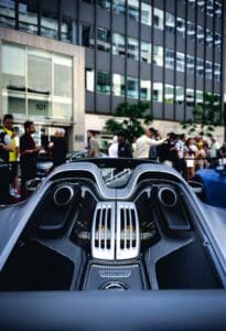 Rear of sleek sports car with vents, engine details; people on city street by office building.