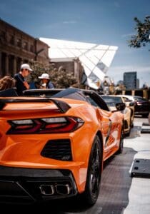 Orange sports car parked on a street with people and modern buildings in the background.