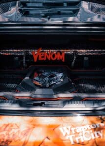 Car trunk with miniature model, red "Venom" sign, orange accents, and mesh background.