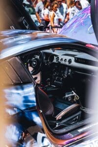 Interior of a sports car with open door, visible dashboard; people in the background.
