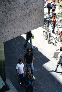 Street scene with pedestrians on a sunny day, bicycles parked nearby, shadows visible.