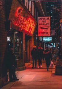 Street scene at night with people near the neon-lit Imperial Pub, Library, and Lounge signs.