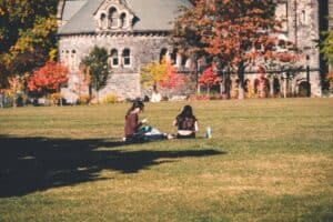 People sitting on grass near a historic stone building with fall trees in the background.