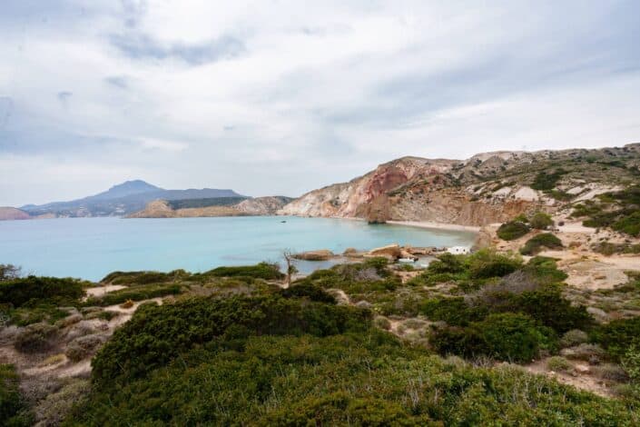A tranquil coastal scene showcases a secluded bay nestled within colorful cliffs, with a mix of lush greenery and light-blue water.