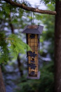 Metal bird feeder with leaf decorations hanging from a tree branch among green leaves.