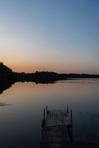Wooden dock extending over calm lake at dusk with trees along the distant shoreline.