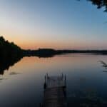 Wooden dock over calm lake with tree-lined shore and sunset sky reflecting on water.