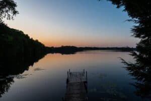 Wooden dock over calm lake with tree-lined shore and sunset sky reflecting on water.